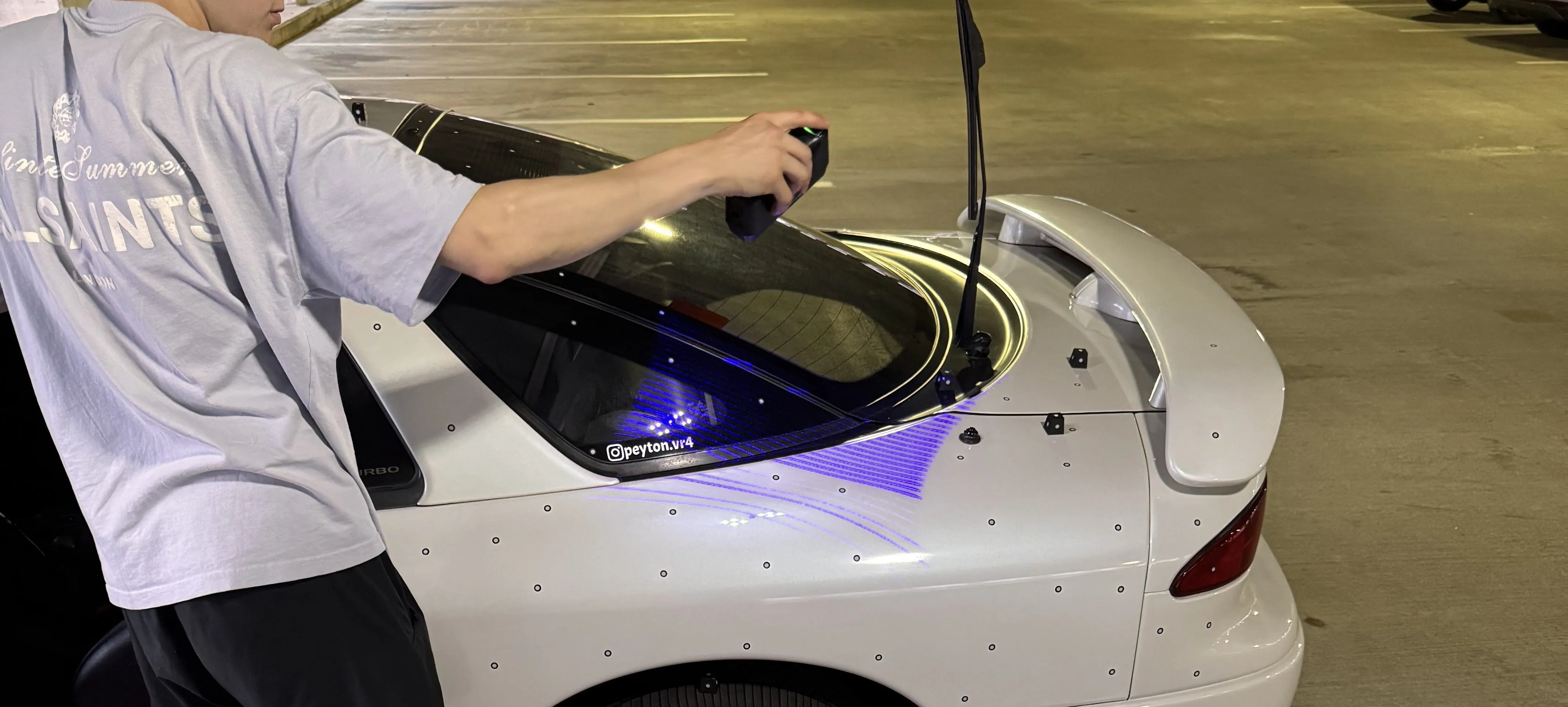 Handheld 3D scanner projecting blue laser lines onto the rear of a white car covered in tracking markers